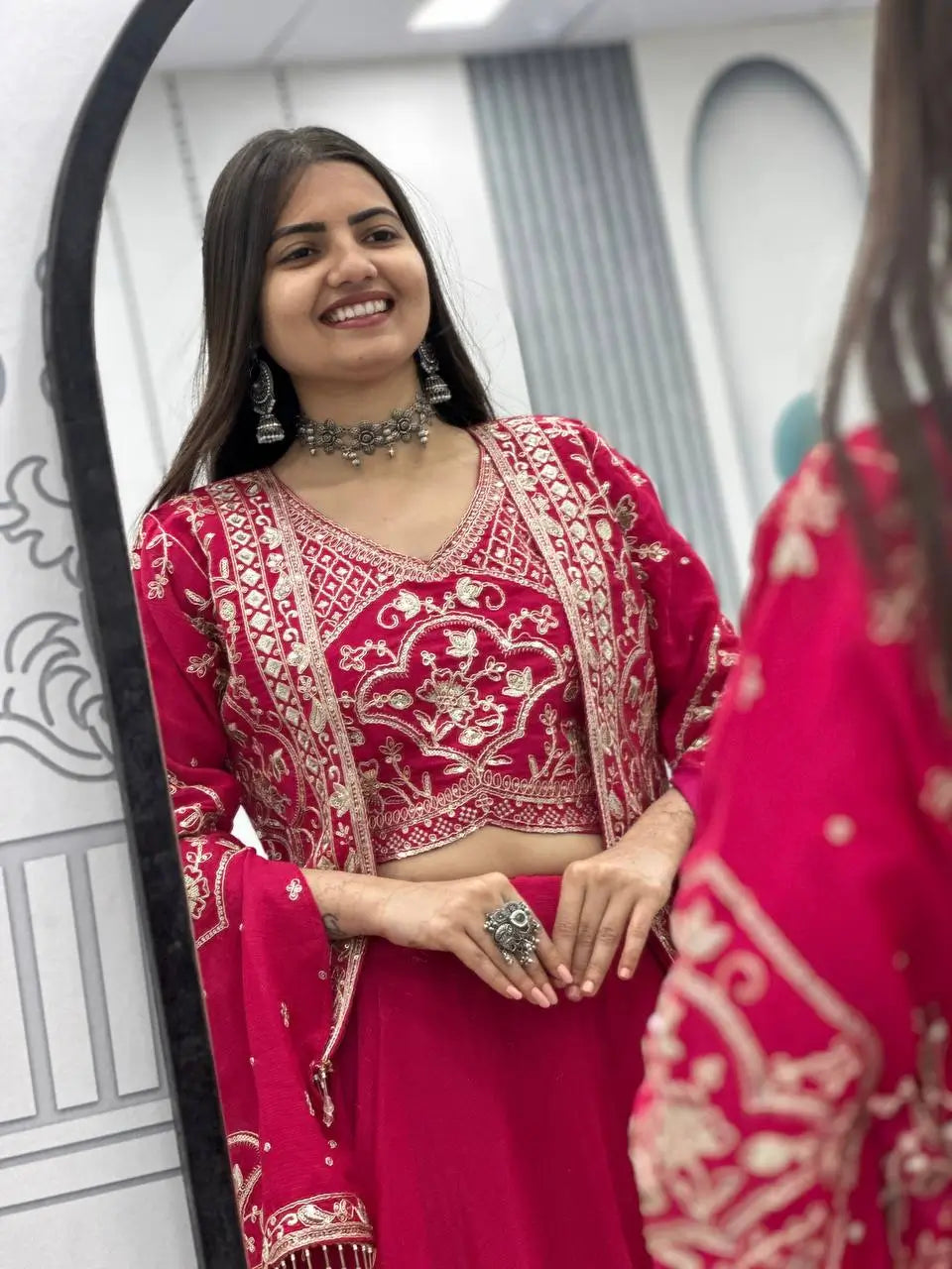 Woman in a red and pink traditional outfit with intricate patterns, standing in front of a mirror.