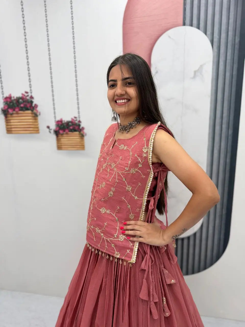 Woman in a pink traditional outfit standing against a decorative wall with plants.