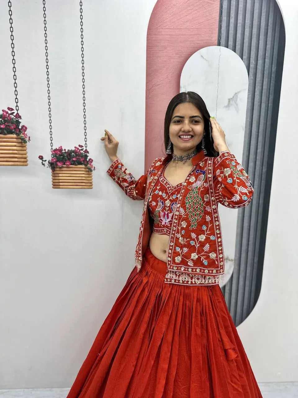 Woman in a red traditional outfit standing in front of a wall with hanging plants and abstract art.