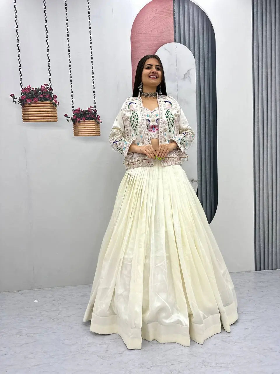 Woman in a floral top and white skirt standing in front of a decorative wall with hanging plants.