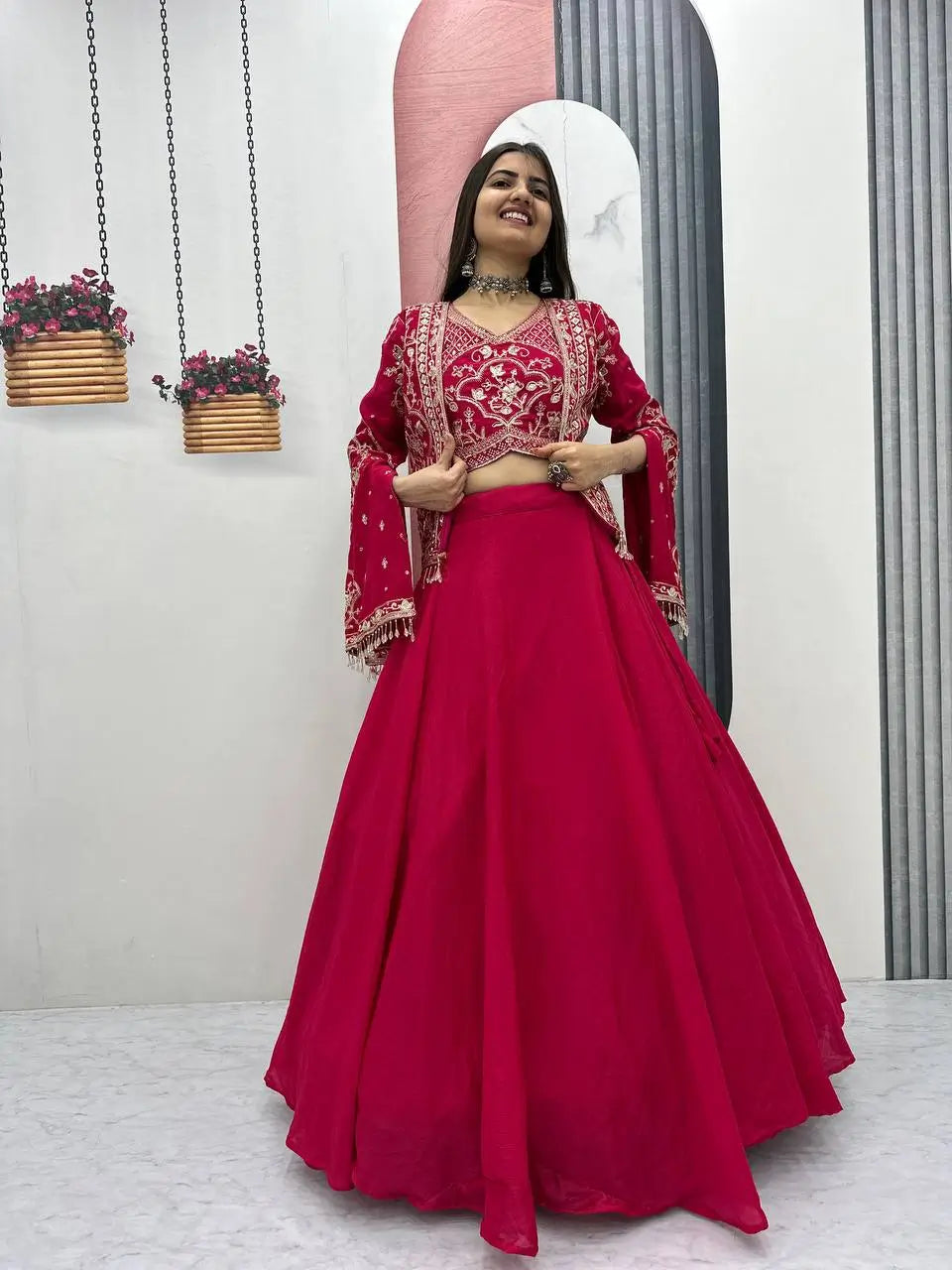 Woman in a red traditional outfit standing in front of a mirror with decorative plants in the background.