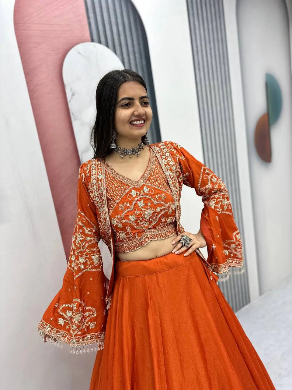 Woman in an orange traditional outfit with intricate patterns, standing against a neutral background.
