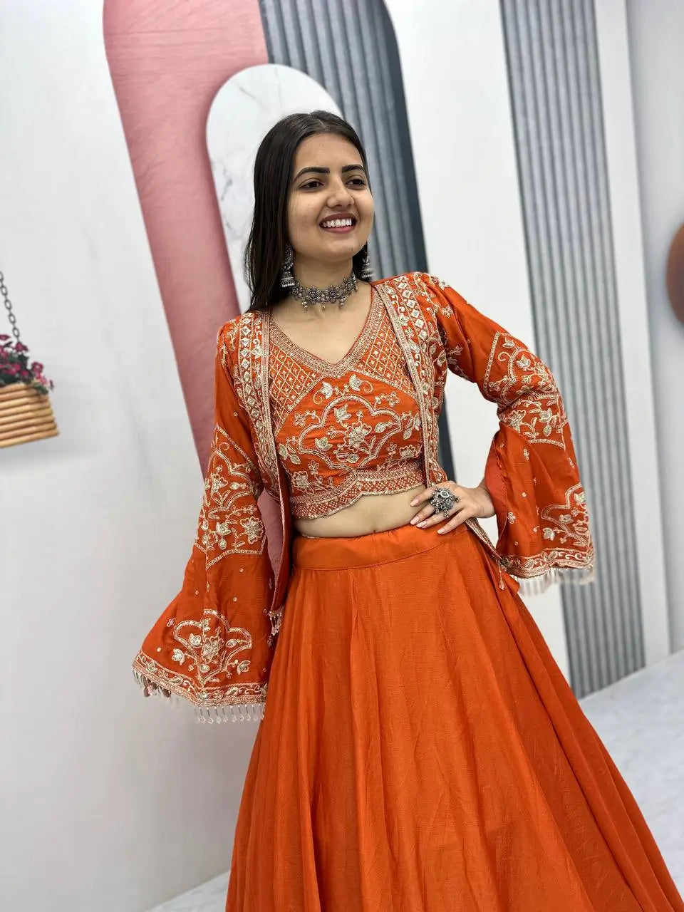 Woman wearing an orange traditional outfit with intricate designs in a minimal indoor setting.
